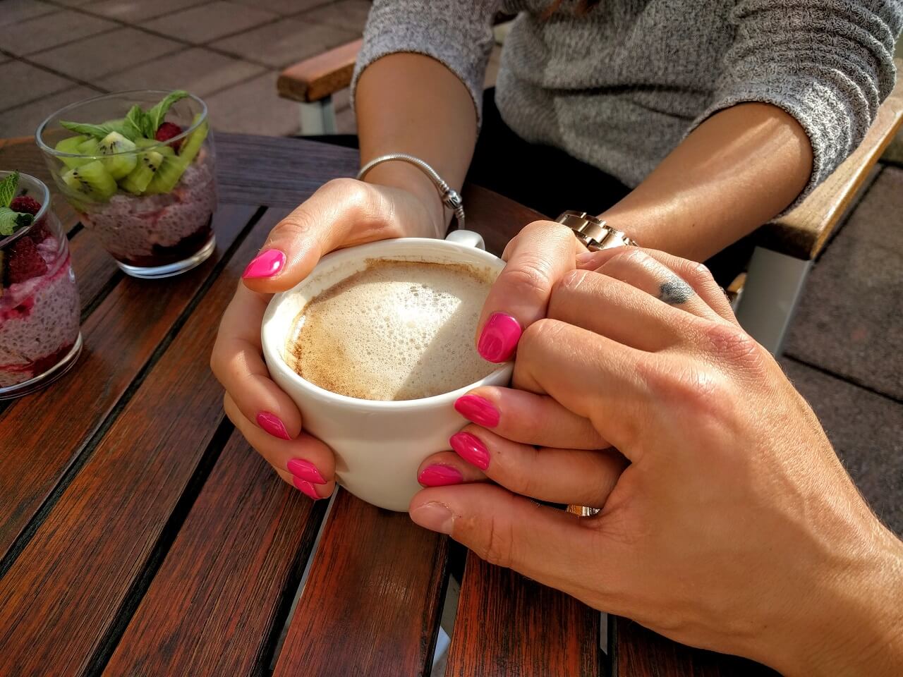 Two people sit at a wooden table. One person holds a cup of coffee in both hands, while the other gently rests their hand on top. Two desserts are on the table.