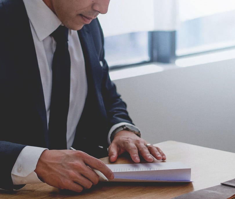 A person in a suit and tie sits at a desk, looking through pages of a document.