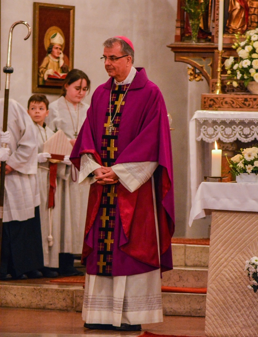 A bishop in purple vestments stands by an altar inside a church, accompanied by altar servers in white robes.