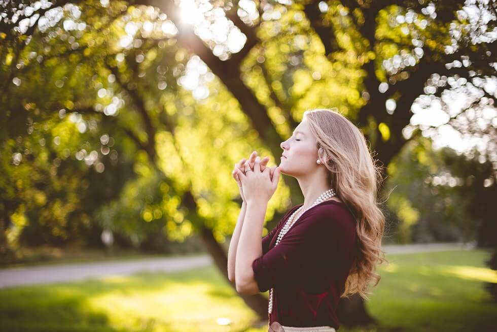 A woman stands outdoors in a sunlit park with her eyes closed and hands clasped, sunlight filtering through trees in the background.