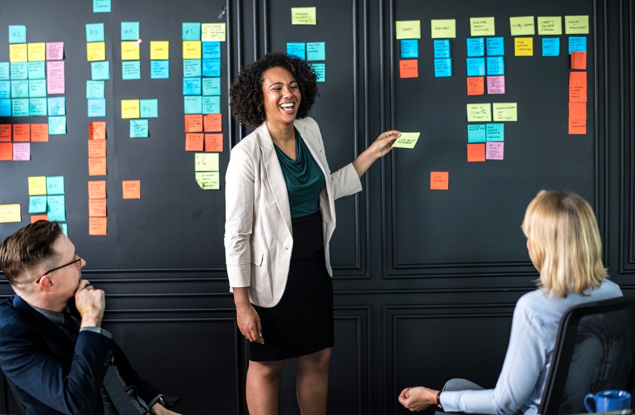 A woman stands and gestures toward a wall covered with colorful sticky notes as two seated colleagues listen attentively.