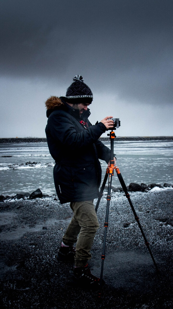 Person dressed in winter clothing photographing an icy shoreline with a camera on a tripod under an overcast sky.