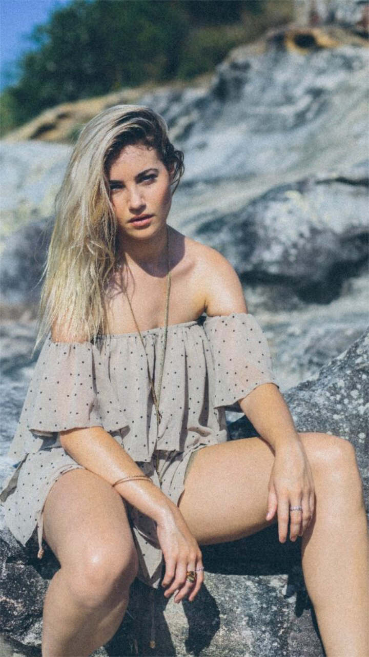 A woman with long blonde hair in an off-shoulder dress sits on outdoor rocks, facing the camera.