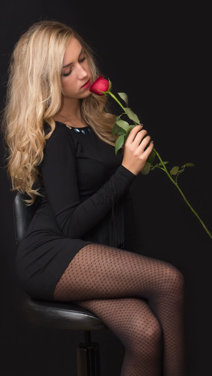 A woman with long blonde hair in a black dress sits on a chair, holding and smelling a red rose against a black background.