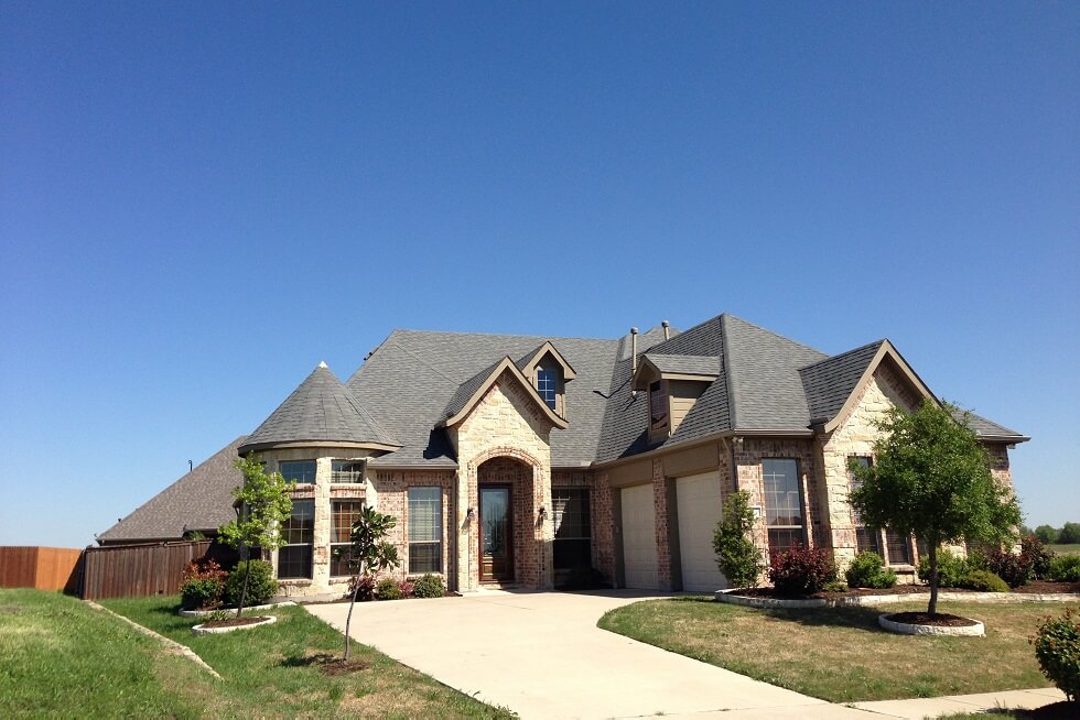 A large suburban brick house with peaked roofs, a two-car garage, and a driveway. Small trees and shrubs are in the front yard under a clear blue sky.