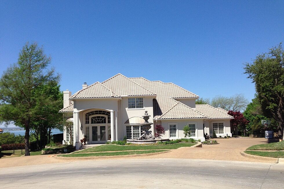 Two-story white house with a tiled roof, circular driveway, and landscaped yard, surrounded by trees on a sunny day.