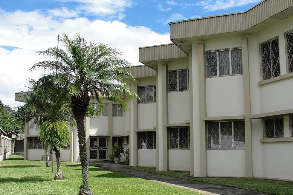 Alt text (refined for WCAG): Two-story cream building with barred windows, palm trees, and a sidewalk on a green lawn under a partly cloudy sky.
