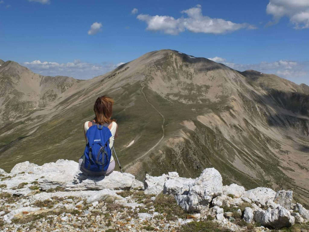 A person wearing a blue backpack sits on rocky ground, facing distant mountains beneath a partly cloudy sky.