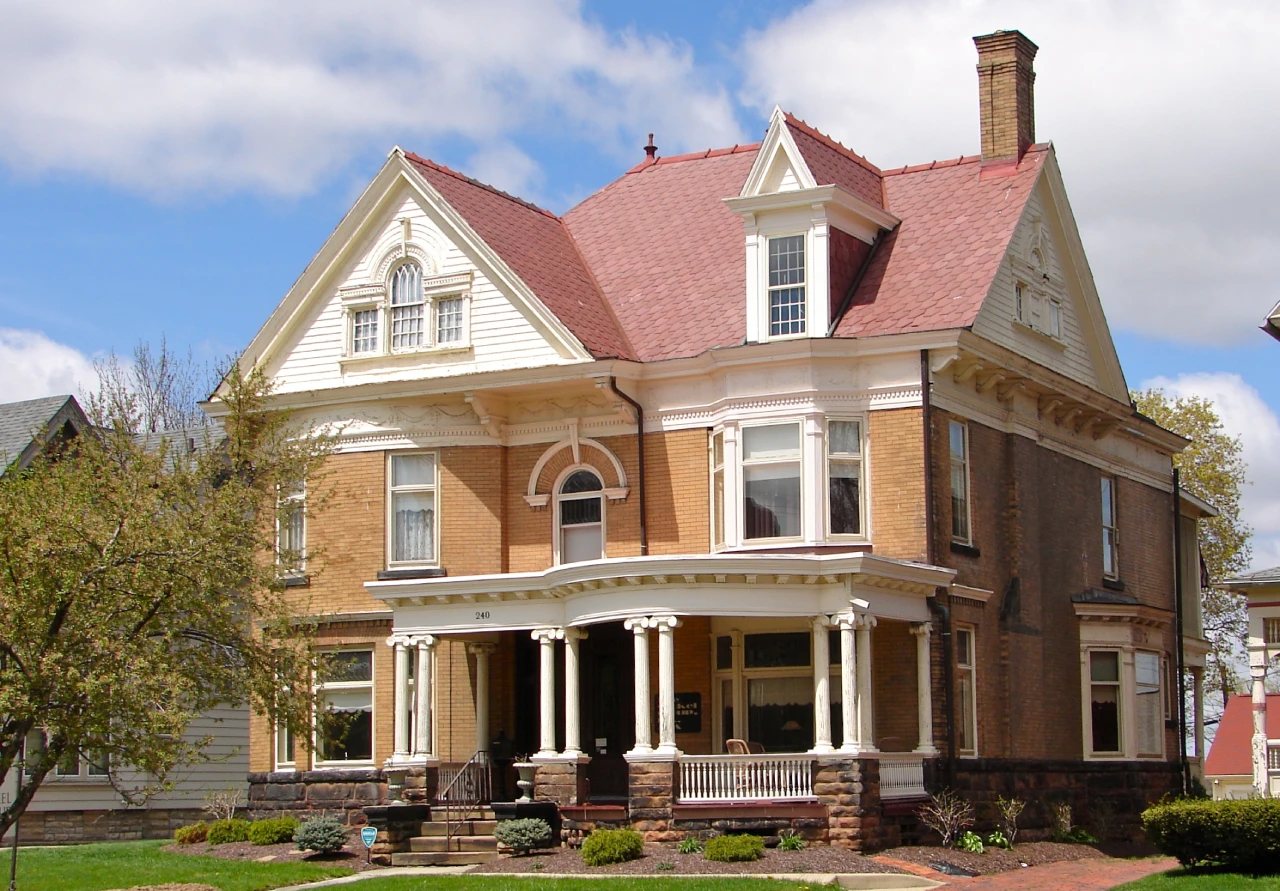 Alt text (WCAG compliant): Victorian-style two-story house with a red roof, decorative trim, large windows, and a wraparound front porch on a landscaped lawn.