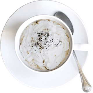 A white cup of a whipped cream-topped beverage garnished with ground black pepper, on a white saucer with a metal spoon.