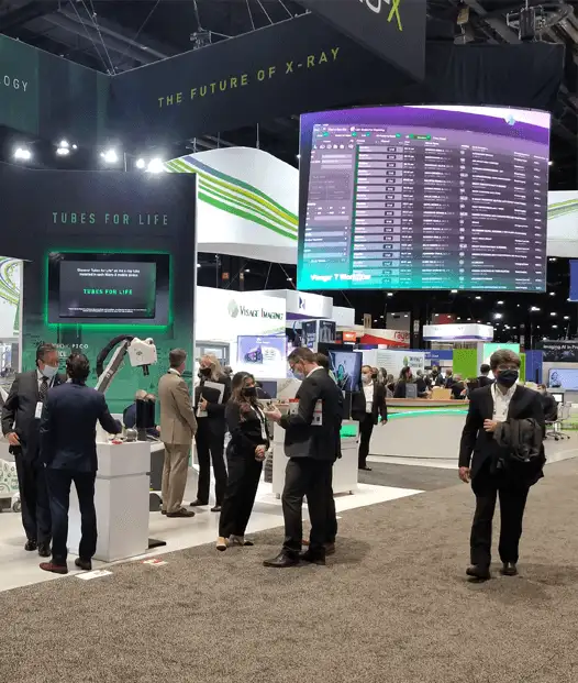 Attendees walk through a trade show with booths, displays, and digital screens showcasing X-ray technology products.