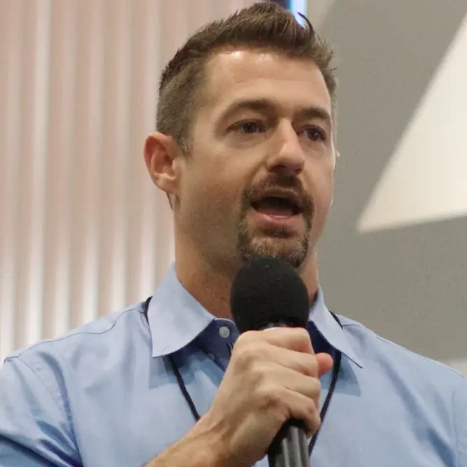 A man in a light blue shirt speaks into a microphone at an indoor event.