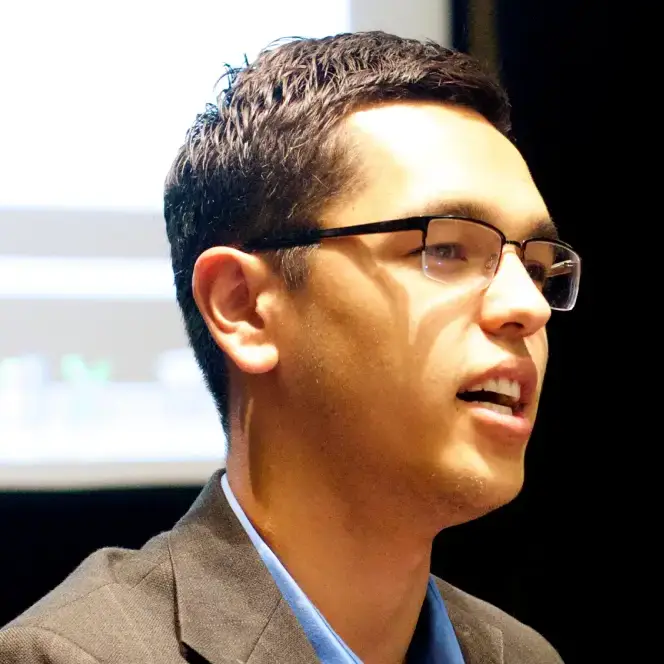 A man with short dark hair and glasses, wearing a blazer and blue shirt, speaks in front of a blurred screen.