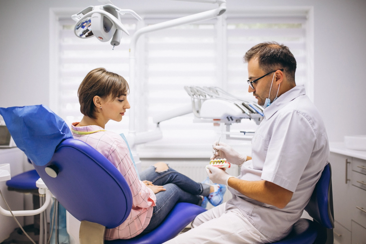 A dentist explains dental care using a tooth model to a patient seated in a dental chair in a well-lit clinic.