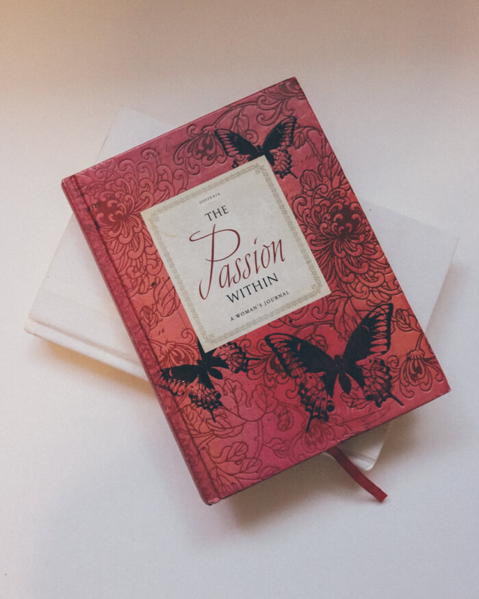 A red hardcover book titled The Passion Within, featuring black butterfly and floral designs on the cover, rests atop a plain white book.
