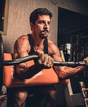 A man in a sleeveless shirt performs seated preacher curls with a barbell in a gym.