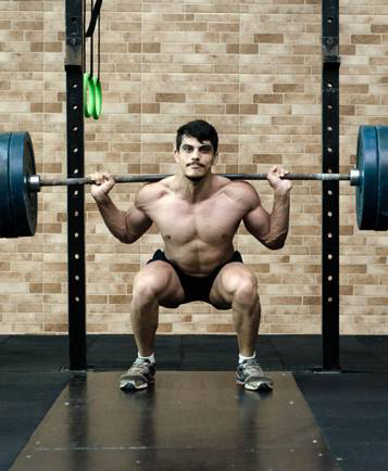 A man with short hair and a mustache performs a barbell back squat in a gym, wearing shorts and sneakers.