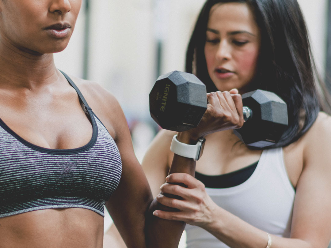 A fitness trainer assists a woman lifting a dumbbell in a gym. Both are wearing athletic clothing.