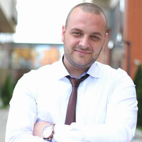 A smiling man with a shaved head and beard, wearing a white shirt and brown tie, stands outdoors with his arms crossed.