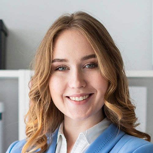A woman with long, wavy blonde hair, wearing a light blue blazer and white shirt, smiles at the camera in a modern office.