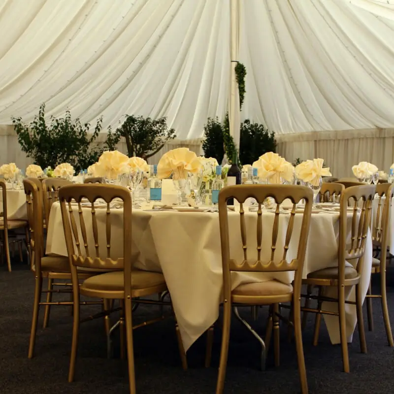 Round tables with beige tablecloths, neatly arranged napkins, and chairs are set for an event inside a draped tent, with greenery visible outside.