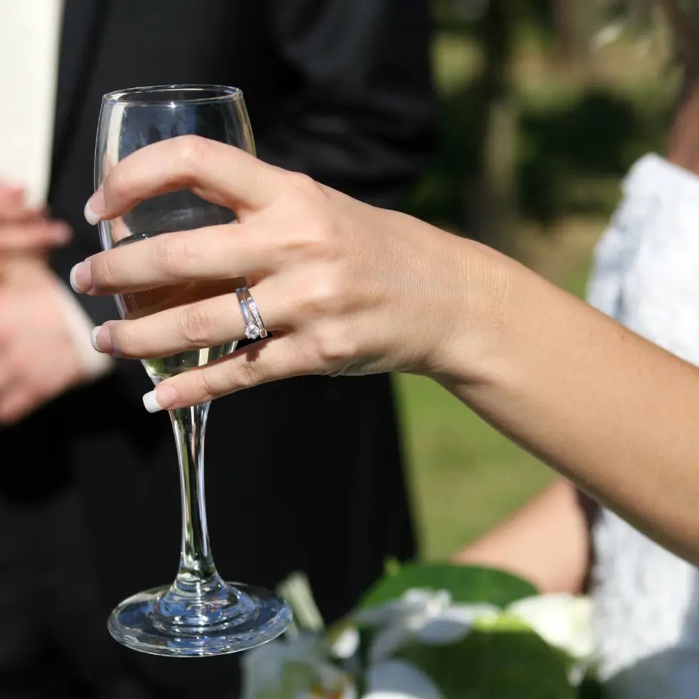 A person in formal attire holds a champagne glass, displaying a wedding ring. A second person in a suit is blurred in the background.