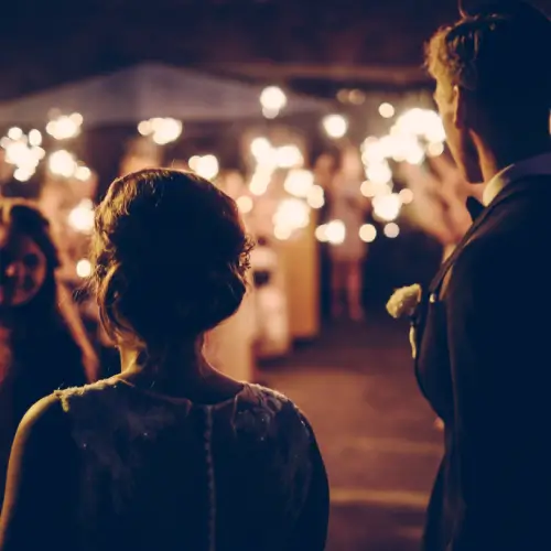A bride and groom stand together at night, facing guests holding sparklers during a wedding celebration.