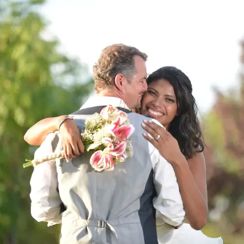 Bride smiling at the camera while hugging her groom outdoors, holding a bouquet of pink lilies behind his back.