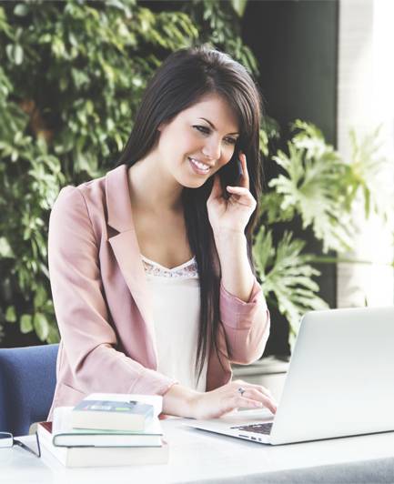 A woman in a pink blazer sits at a desk, talking on the phone and using a laptop, with books and plants in the background.