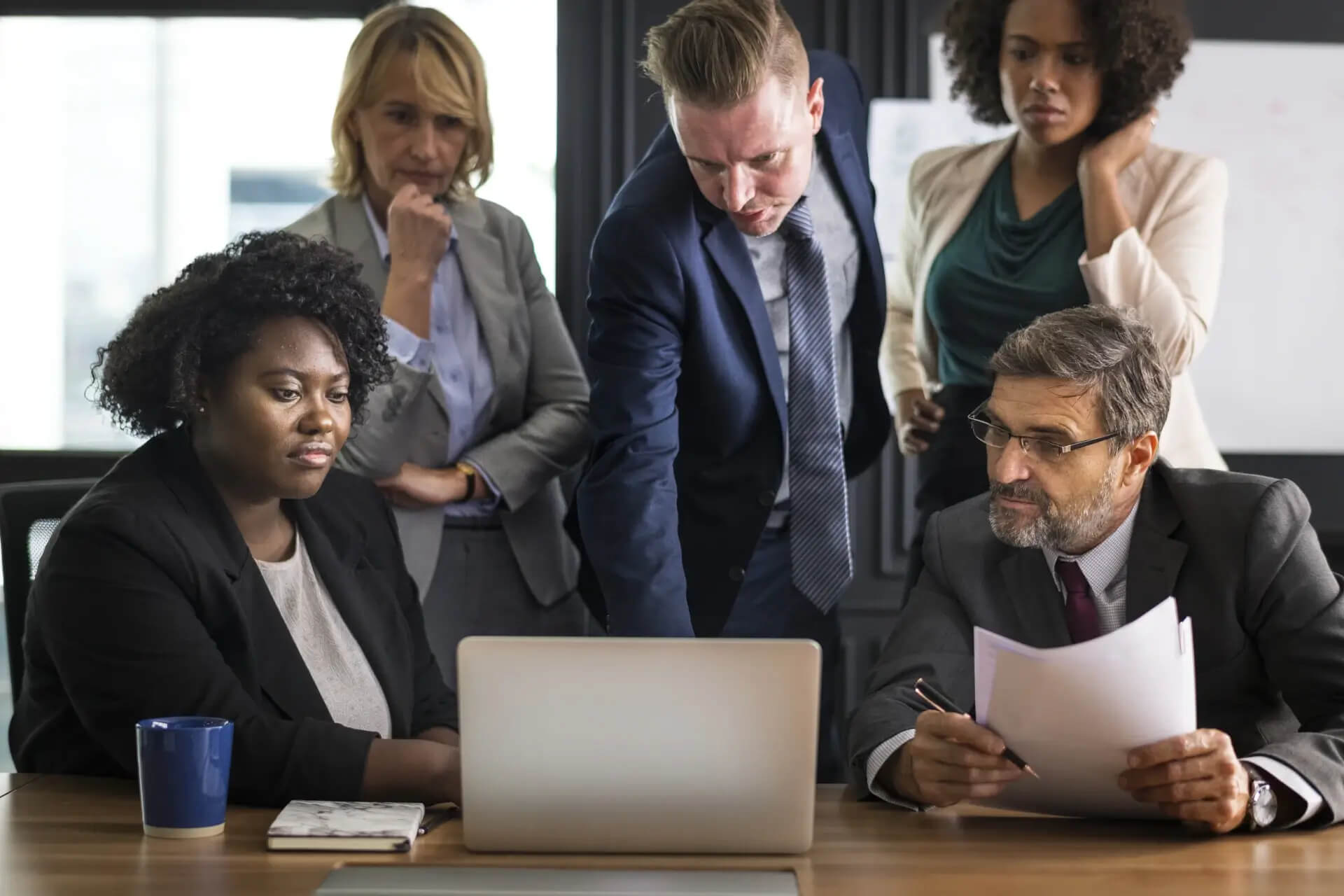 Five business professionals sit around a table with documents and a coffee mug, looking at a laptop and having a serious discussion.