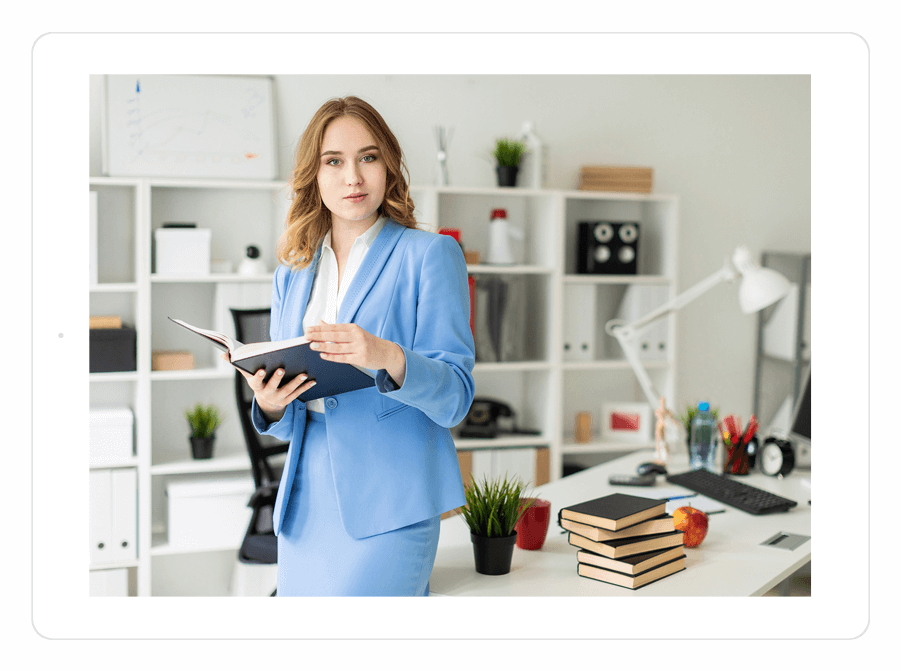 A woman in a blue suit stands in an office holding an open book. Books, a desk, and shelves with office items are visible in the background.