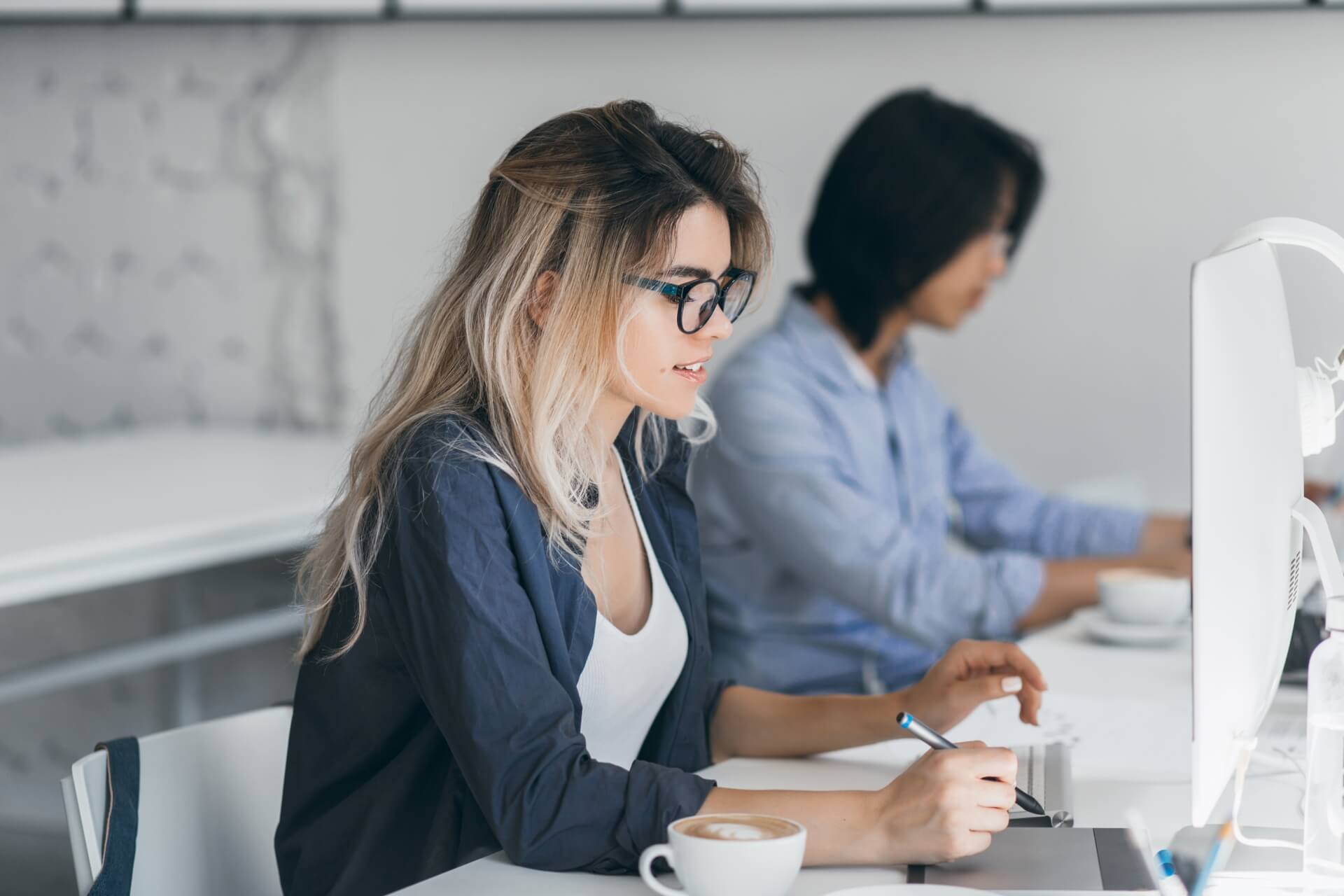 A woman sits at a desk in an office, holding a pen and looking at her computer monitor with a coffee cup beside her. Another person works at a desk in the background.