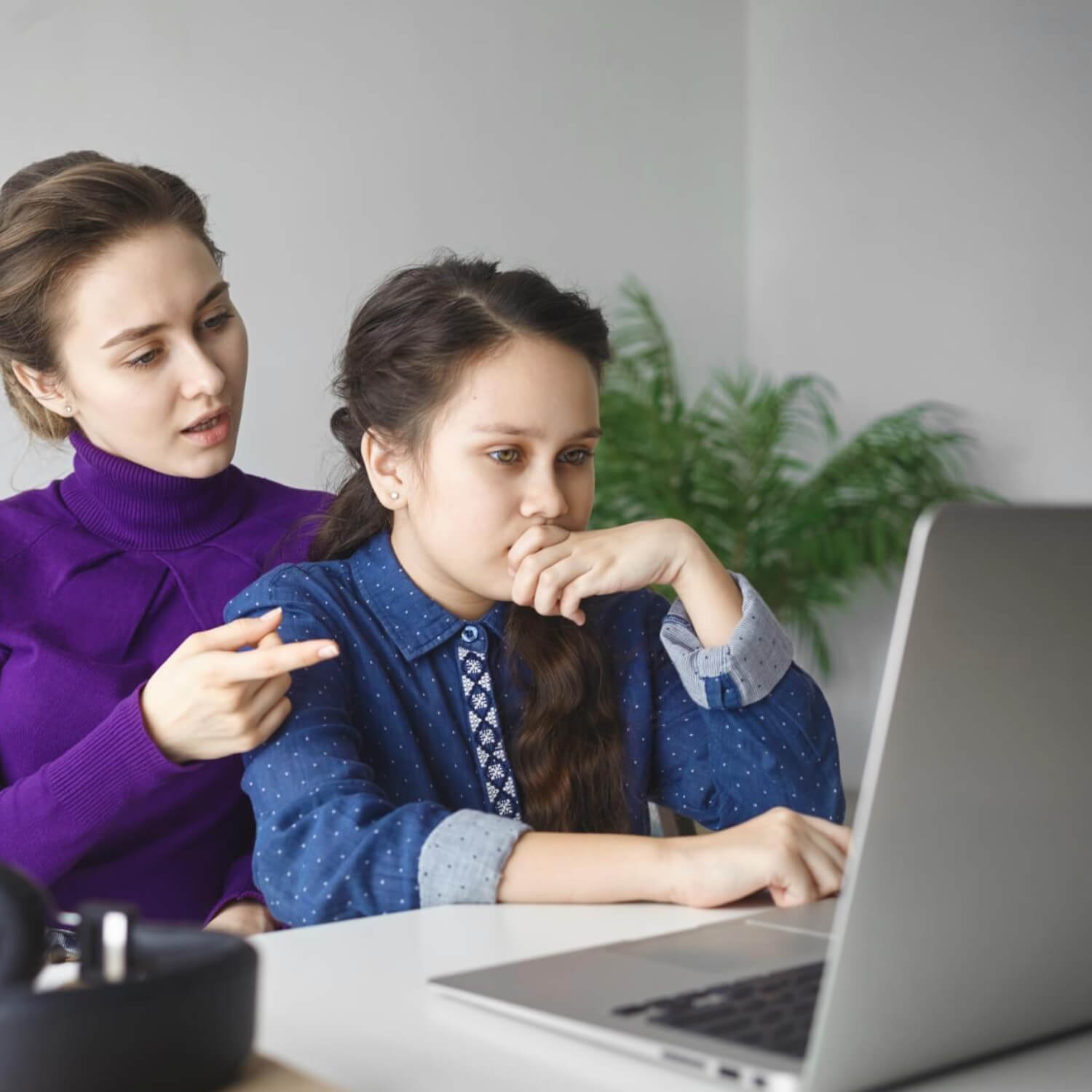 A woman points at a laptop screen while a young girl watches thoughtfully; both are seated together at an indoor table.