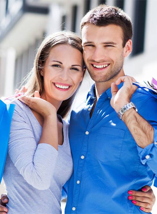 A man and woman smile while standing close together outdoors, each holding shopping bags over their shoulders.