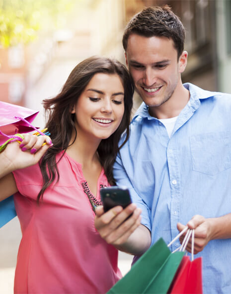 Two people, a woman and a man, stand outdoors holding shopping bags and smiling as they look at a smartphone together.