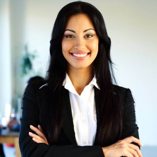 A woman with long dark hair, wearing a black blazer over a white shirt, stands indoors with her arms crossed and smiles at the camera.