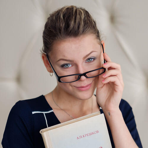 A woman wearing glasses holds a book and looks at the camera against a plain background.
