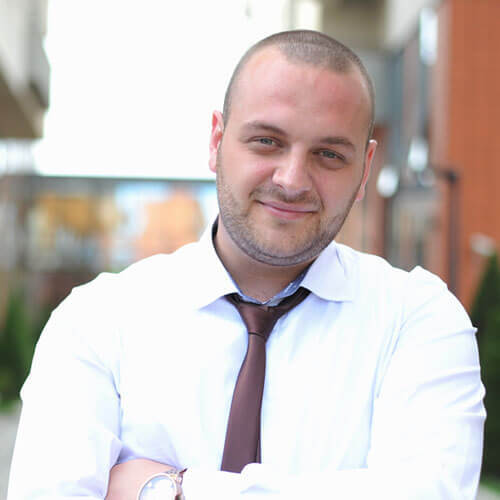 A man with a shaved head and light beard stands outdoors with his arms crossed, wearing a white shirt and brown tie, smiling slightly.