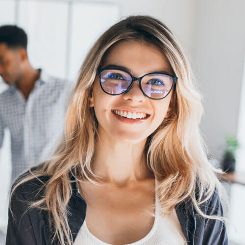 A woman with long blonde hair and glasses smiles at the camera indoors; a man stands blurred in the background.