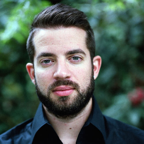 A man with short brown hair and a beard, wearing a black collared shirt, stands outside with blurred green foliage in the background.