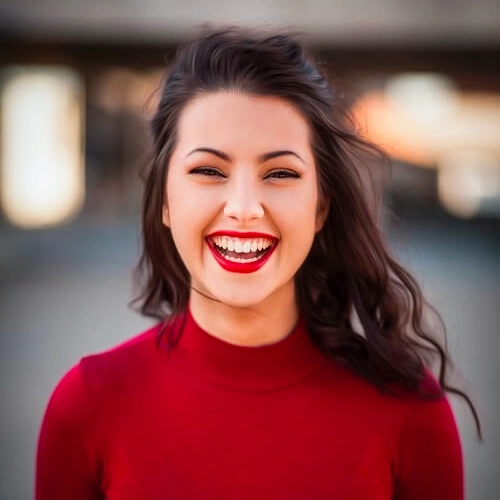 A woman with long brown hair, wearing a red top, smiles outdoors in front of a blurred background.